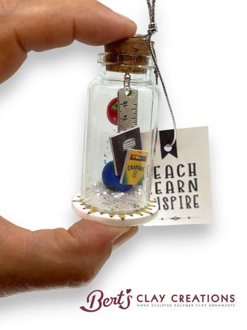 Hand holding a small glass jar with a cork lid containing teacher or education decorative items, with 'Bert's Clay Creations' branding.